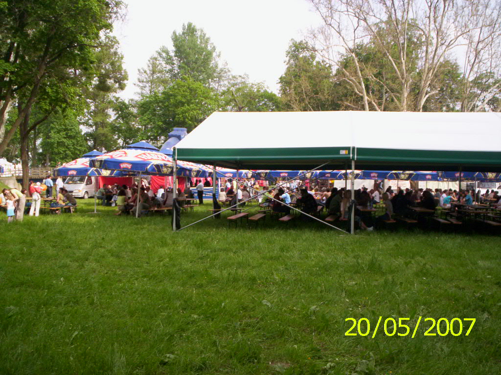 Widok z zewnątrz na imprezę plenerową z białym namiotem z zielonym obramowaniem i ławami piwnymi pod spodem, w tle widoczne parasole piwne i bawiący się ludzie. Data 20/05/2007 widoczna w prawym...